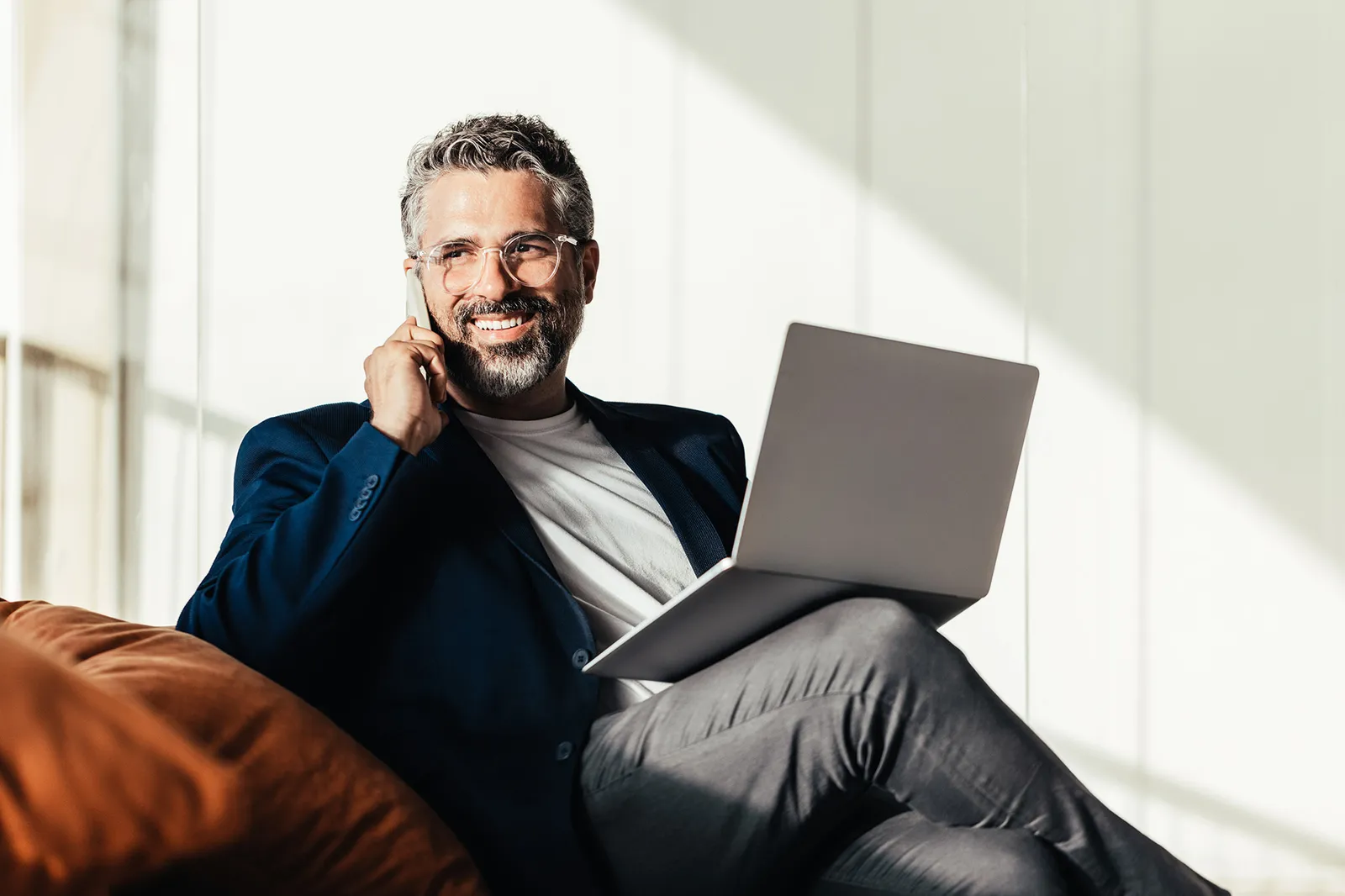 Un uomo è seduto sul divano con un computer portatile e parla al telefono.
