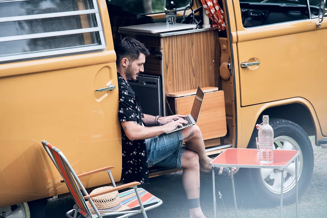 Ein Mann sitzt in der offenen Tür eines Campingbusses und arbeitet mit einem Laptop.