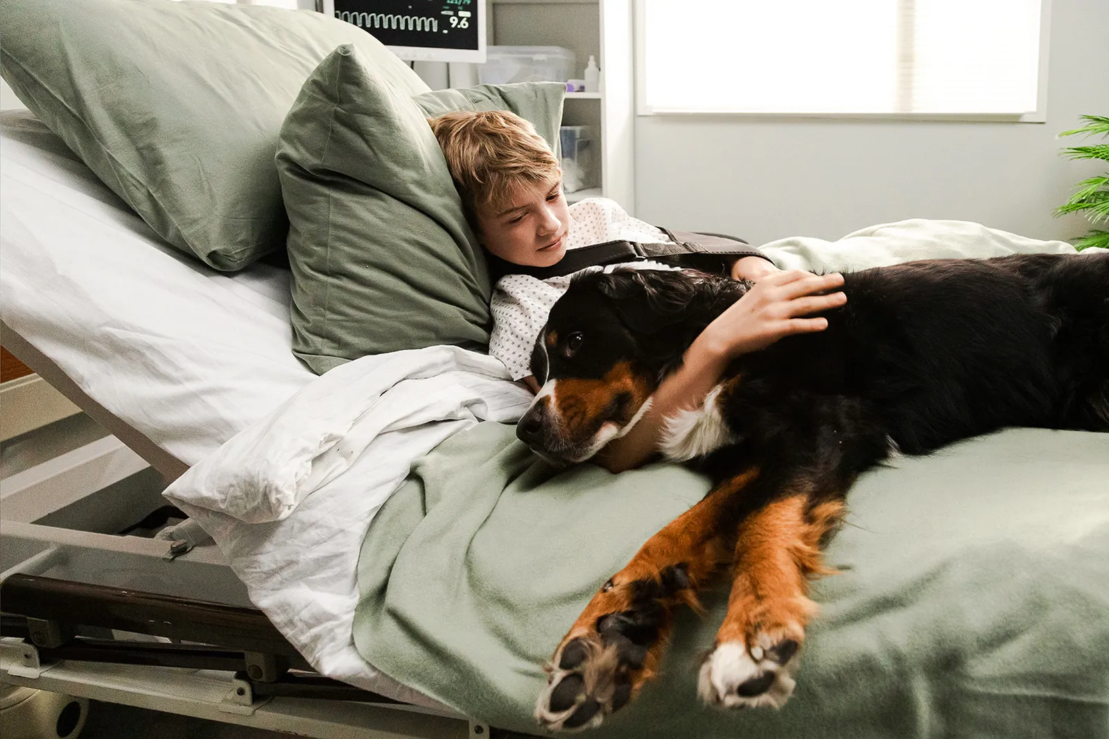Un ragazzo è sdraiato in un letto d’ospedale e accarezza un cane.
