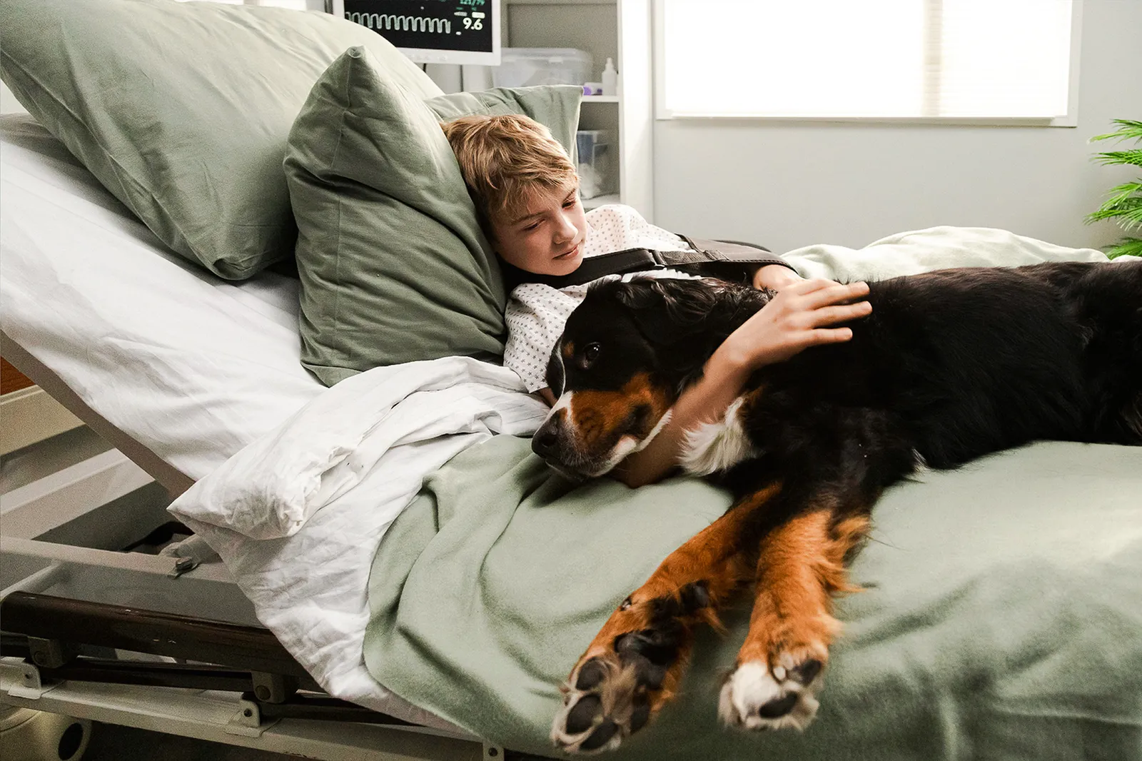 Un ragazzo è sdraiato in un letto d’ospedale e accarezza un cane.
