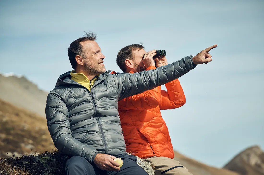 Zwei Männer sitzen in den Bergen, einer schaut durch ein Fernglas und der andere zeigt in die Ferne.