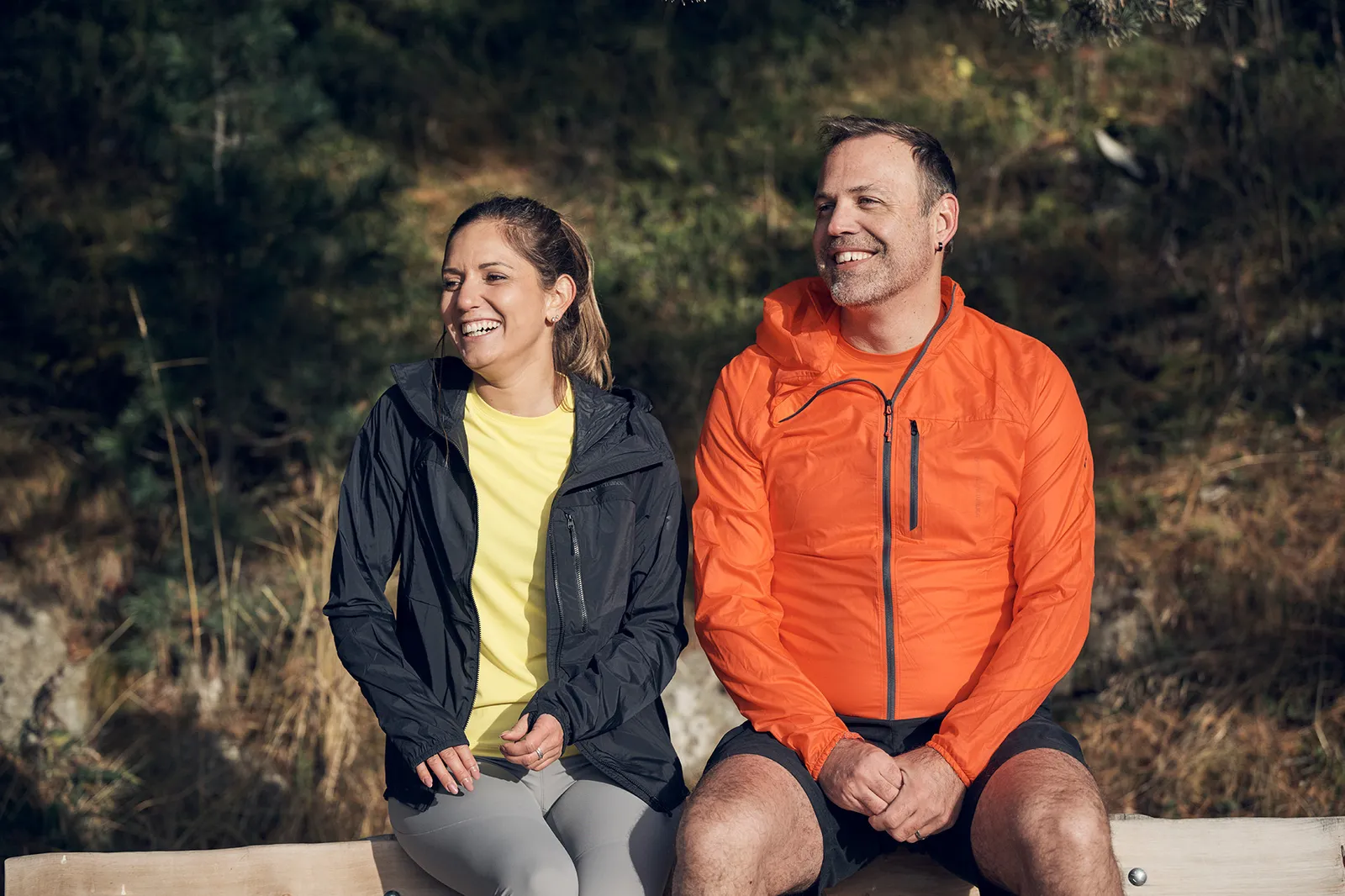 Una donna e un uomo in abbigliamento sportivo sono seduti su una panchina di legno nella natura e sorridono.
