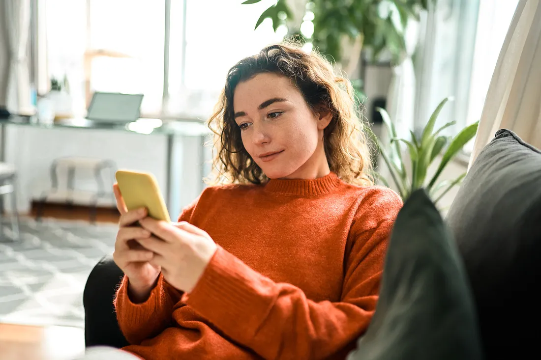 Una donna è seduta sul divano a casa e guarda il suo smartphone.