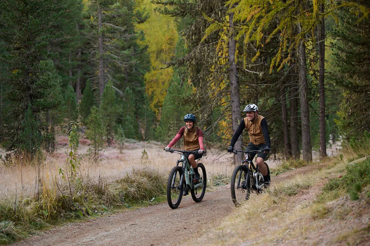 Zwei Radfahrer in braunen Westen fahren auf einem Waldweg umgeben von Nadelbäumen und herbstlicher Vegetation