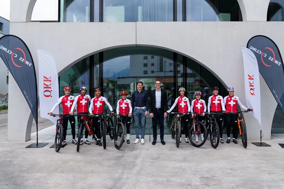 Team von Radfahrern in roten Trikots mit Schweizer Kreuz vor dem Hauptsitz von ÖKK in Landquart, flankiert von Fahnen