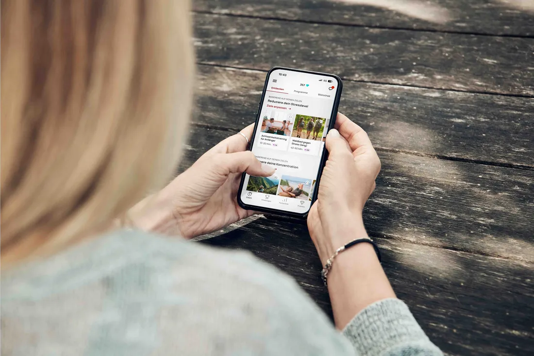 Person nutzt eine Gesundheits-App auf dem Smartphone und scrollt durch Programme auf einem Holztisch.