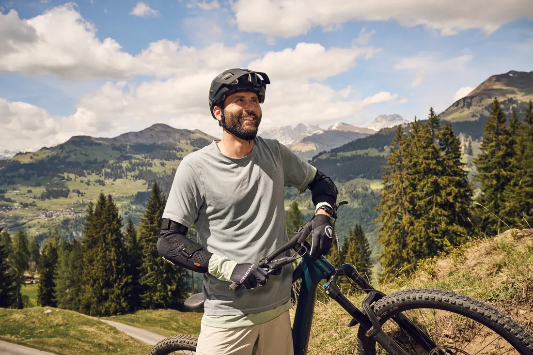 Una persona con una mountain bike si trova su un sentiero di montagna con vista sul paesaggio alpino circostante.