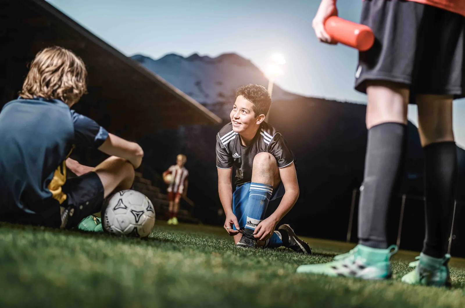 Giovani calciatori fanno una pausa sul campo; uno si allaccia le scarpe mentre gli altri sono seduti e in piedi accanto.