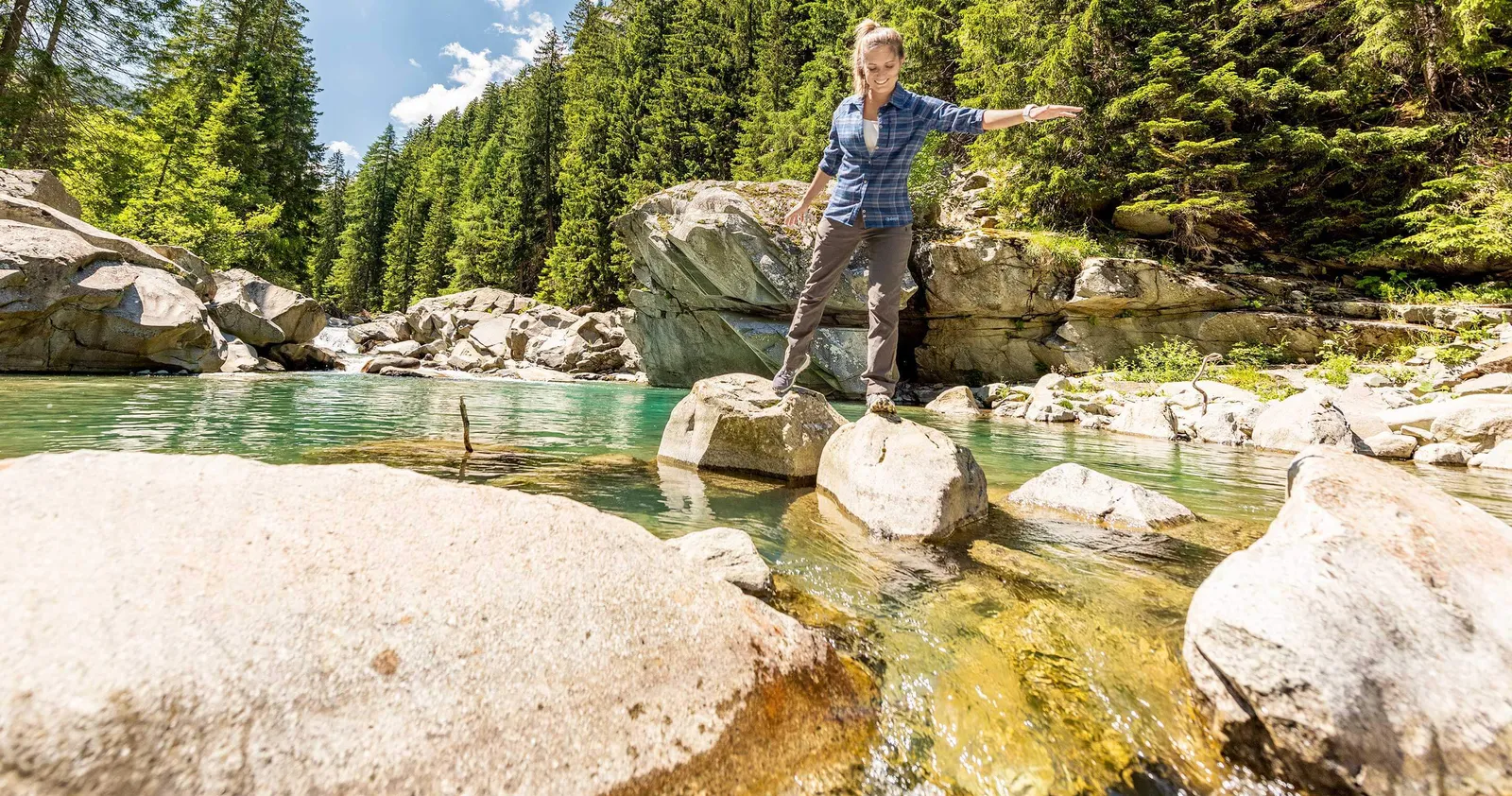 Frau balanciert auf Felsen in einem klaren Gebirgsbach, umgeben von Nadelwald und blauem Himmel