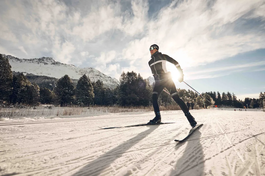 Person beim Langlaufen auf einer sonnigen Loipe vor verschneiten Bergen und Wald.