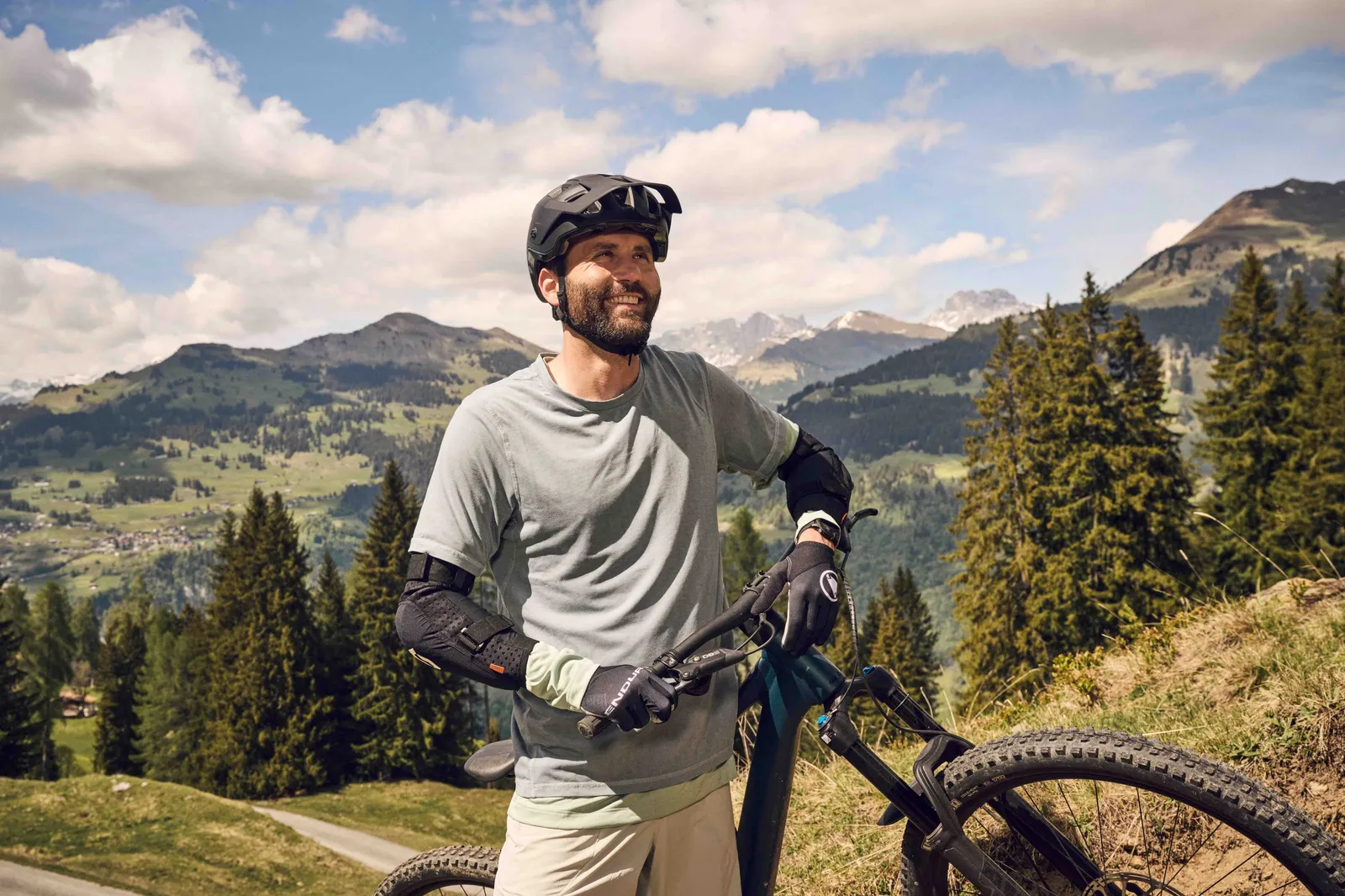 Una persona con una mountain bike e dispositivi di protezione si trova su un sentiero di montagna davanti a un vasto paesaggio alpino con boschi e cime montuose.