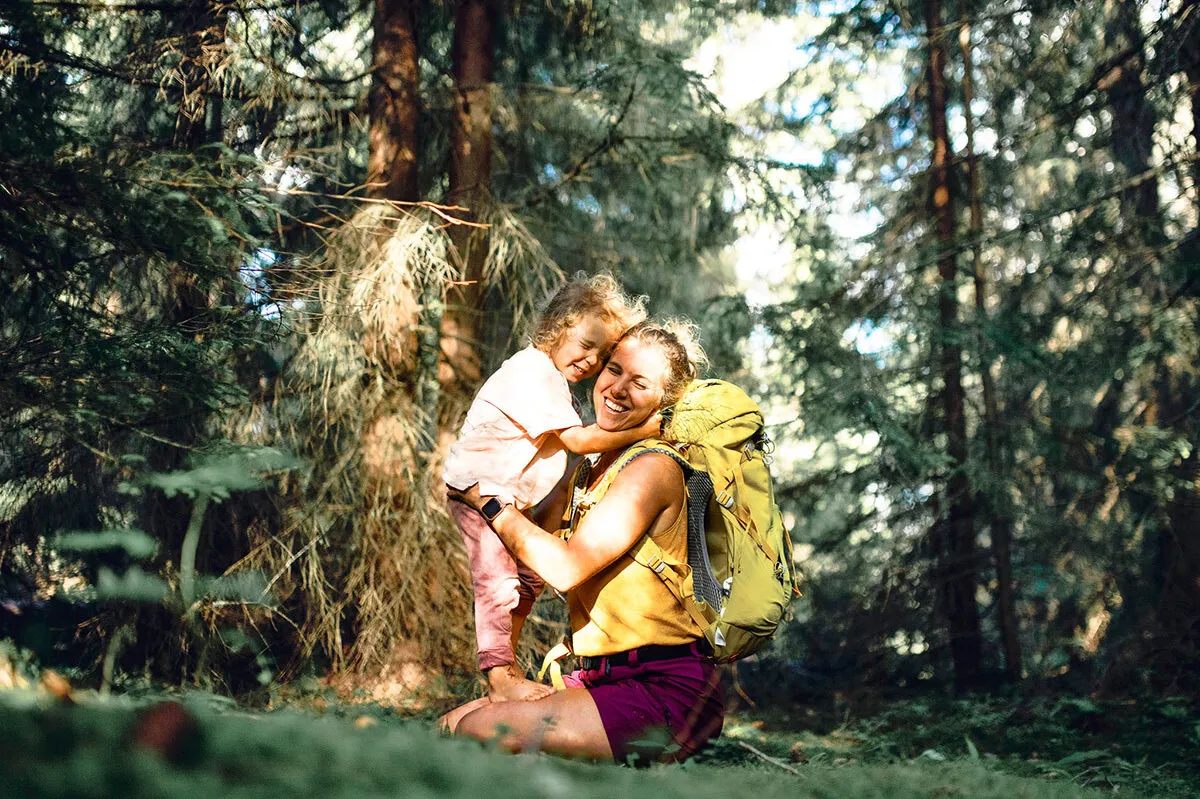Una donna abbraccia sua figlia durante un’escursione nel bosco; entrambe sorridono radiosamente.