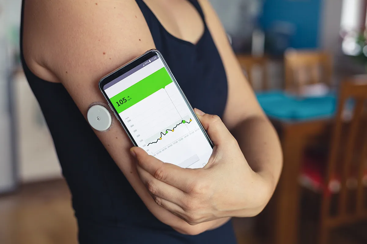 Une personne porte un capteur de glucose sur le bras et tient un smartphone affichant une courbe glycémique autour de 105 mg/dl.