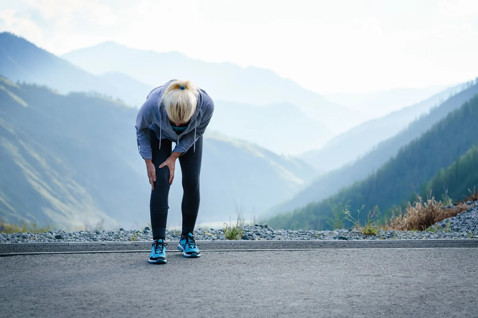 Eine joggende Frau beugt sich erschöpft nach vorne und hält sich schmerzhaft das Knie vor einer Berglandschaft.