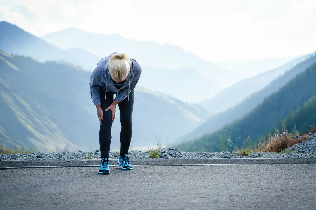 Eine joggende Frau beugt sich erschöpft nach vorne und hält sich schmerzhaft das Knie vor einer Berglandschaft.