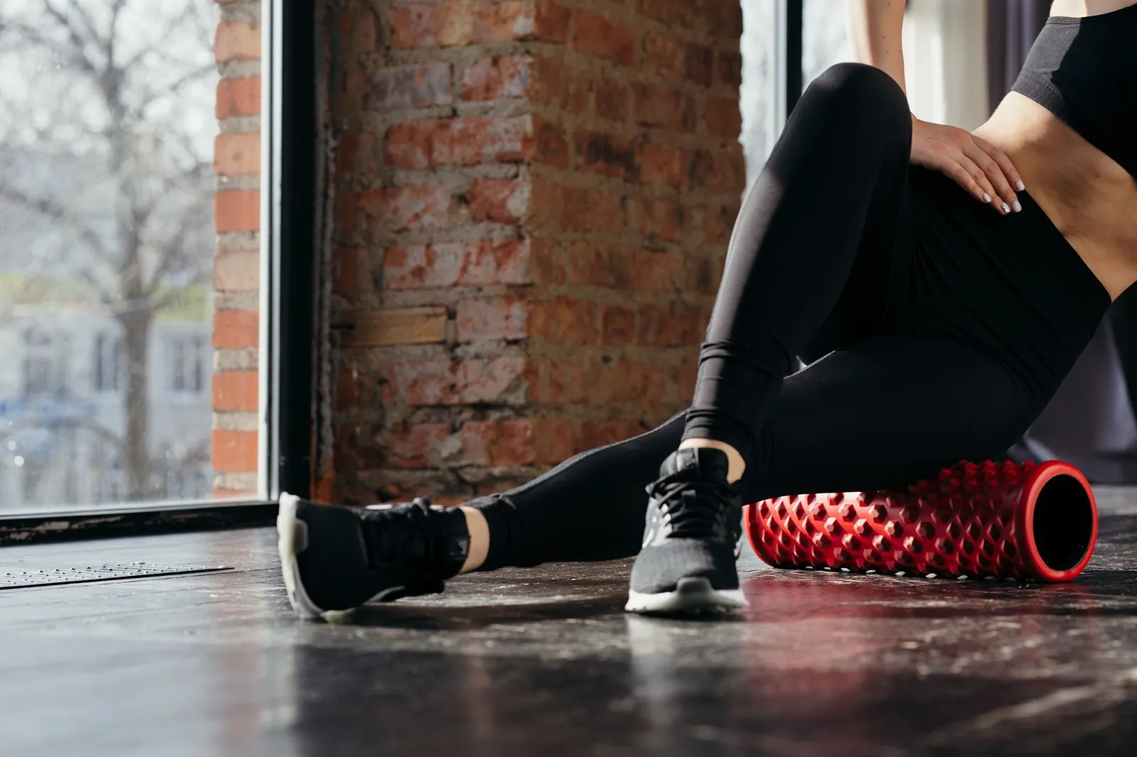 Persona in abbigliamento sportivo utilizza un rullo massaggiante rosso per il recupero muscolare sul pavimento di uno spazio di allenamento luminoso.