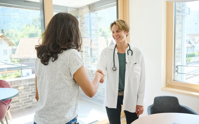 Ärztin mit weissem Kittel und Stethoskop schüttelt einer Frau mit dunklen, lockigen Haaren die Hand in einem hellen Behandlungszimmer.