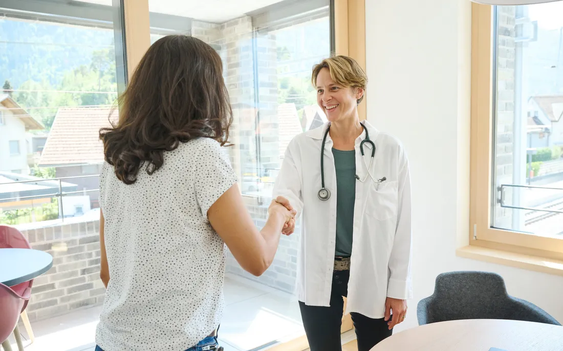 Ärztin mit weissem Kittel und Stethoskop schüttelt einer Frau mit dunklen, lockigen Haaren die Hand in einem hellen Behandlungszimmer.