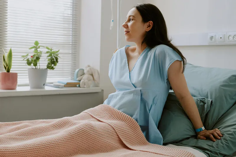 Frau im hellblauen Krankenhaushemd sitzt auf einem Bett, mit rosa gestrickter Decke bedeckt, neben einem Fenster mit Pflanzen.