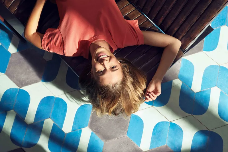 Eine Frau liegt entspannt auf einem braunen Ledersessel und lächelt in die Kamera. Unter ihr ist ein auffälliger Boden mit blau-weissem Muster.