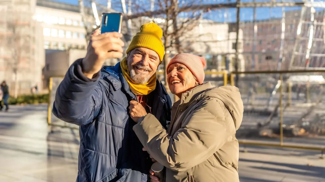 Lächelndes älteres Paar macht ein Selfie im Freien an einem sonnigen Tag, trägt Winterjacken und bunte Strickmützen.