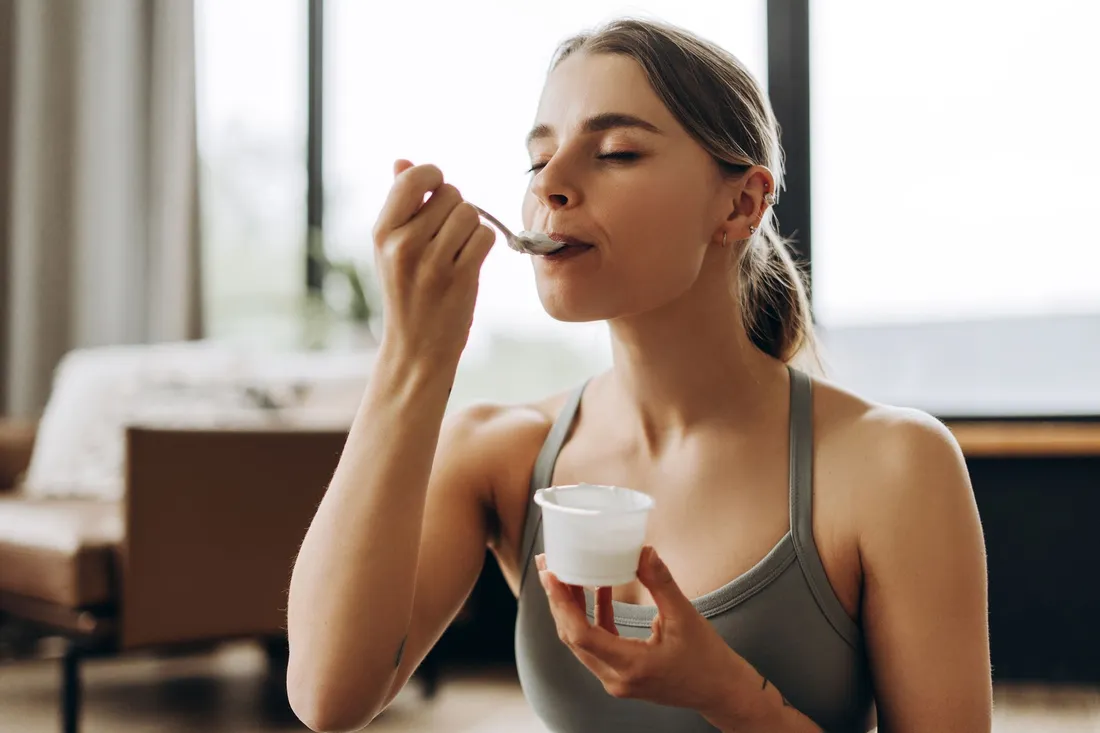 Eine Frau isst mit geschlossenen Augen genüsslich einen Löffel Joghurt in einer hellen, gemütlichen Umgebung.