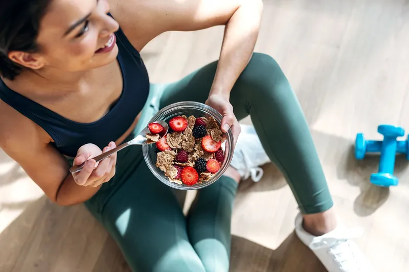 Person in Sportkleidung sitzt auf dem Boden und isst ein Müesli mit Beeren neben einer Hantel.