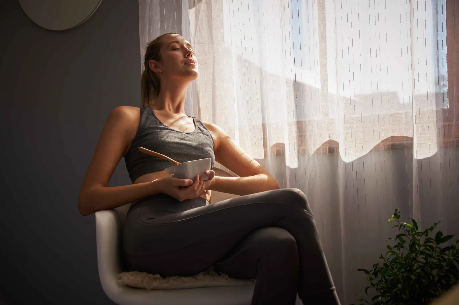 Frau in Sportkleidung sitzt am Fenster auf einem Stuhl, hält eine Schüssel Suppe und lässt sich von der Sonne blenden.