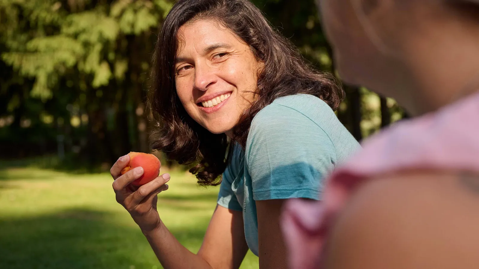 Frau mit braunen Haaren und blauem T-Shirt lächelt und hält einen Apfel im Park