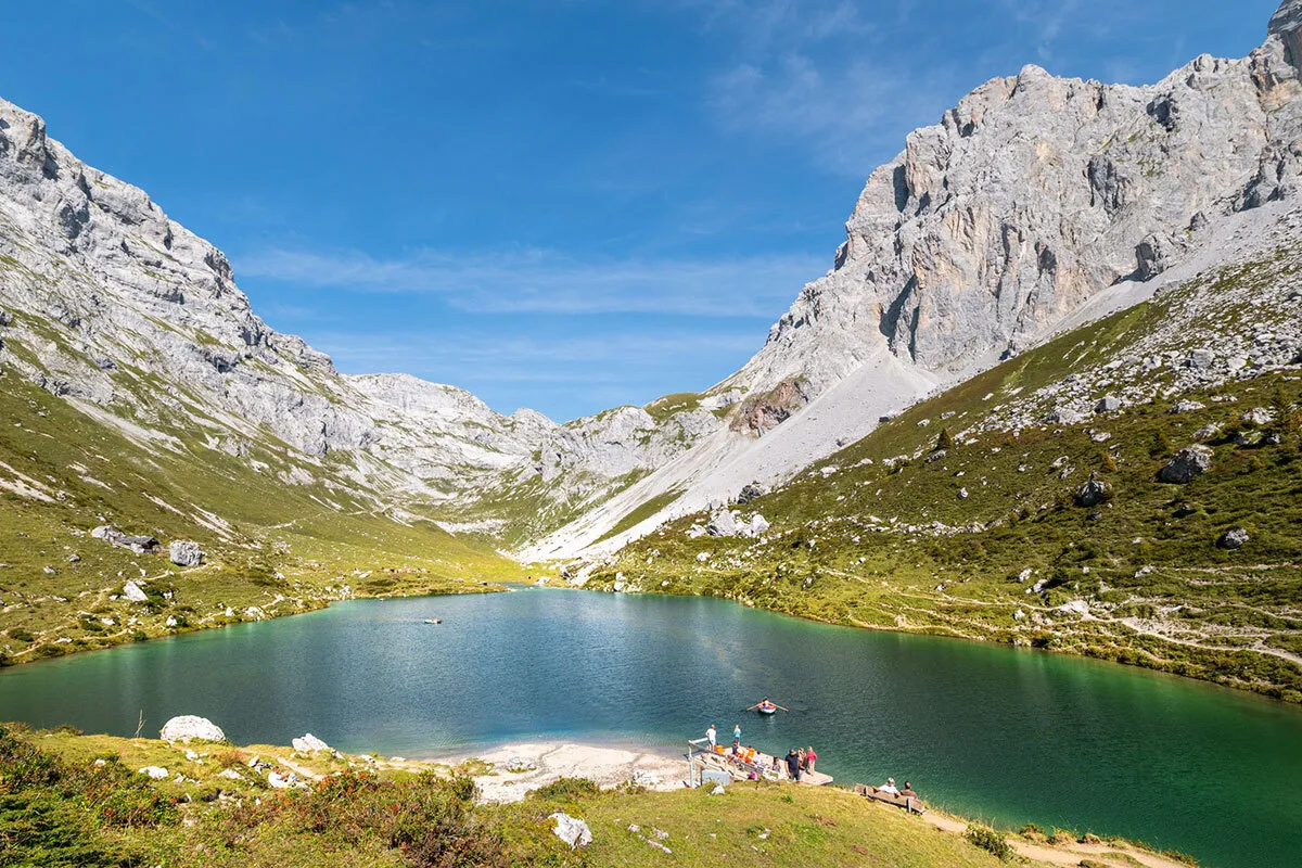 Der Partnunsee im Prättigau, umgeben von hohen Bergen und grünem Gras, mit Menschen am Ufer und Booten auf dem Wasser.