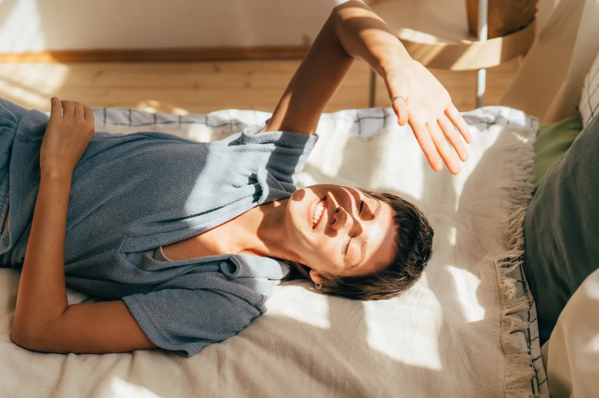 Una persona è distesa su un letto o un divano e solleva una mano per schermarsi dalla luce del sole.