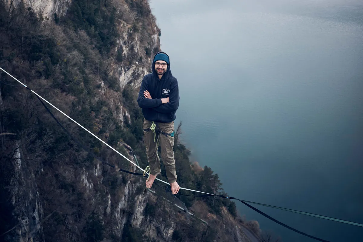 Thomas Schneebeli posiert kühn auf der Slackline hoch über einem malerischen See, umgeben von rauer Natur.