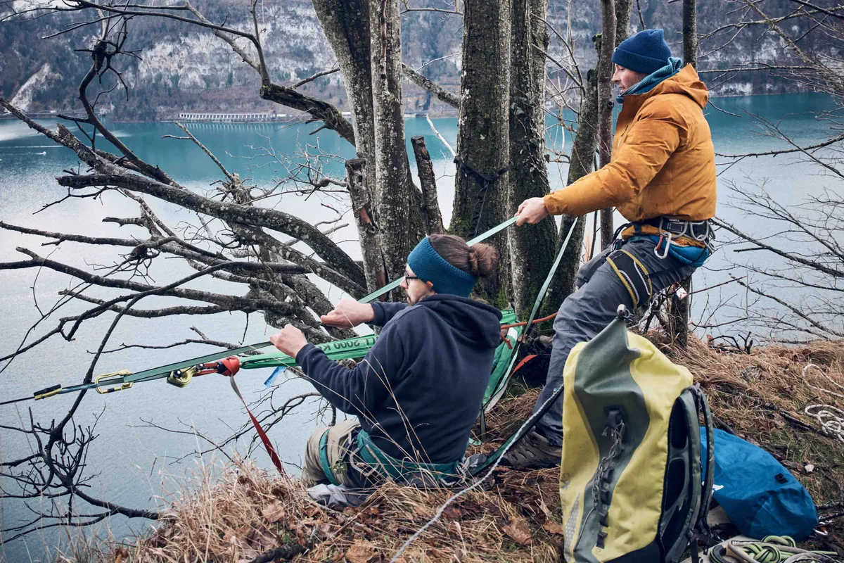 Zwei Männer bereiten sich sorgfältig vor, um eine Slackline über einem See zu spannen, während sie auf einem Felsvorsprung stehen. Sie überprüfen Sicherheitsvorkehrungen, bevor sie sich der Herausforderung stellen.