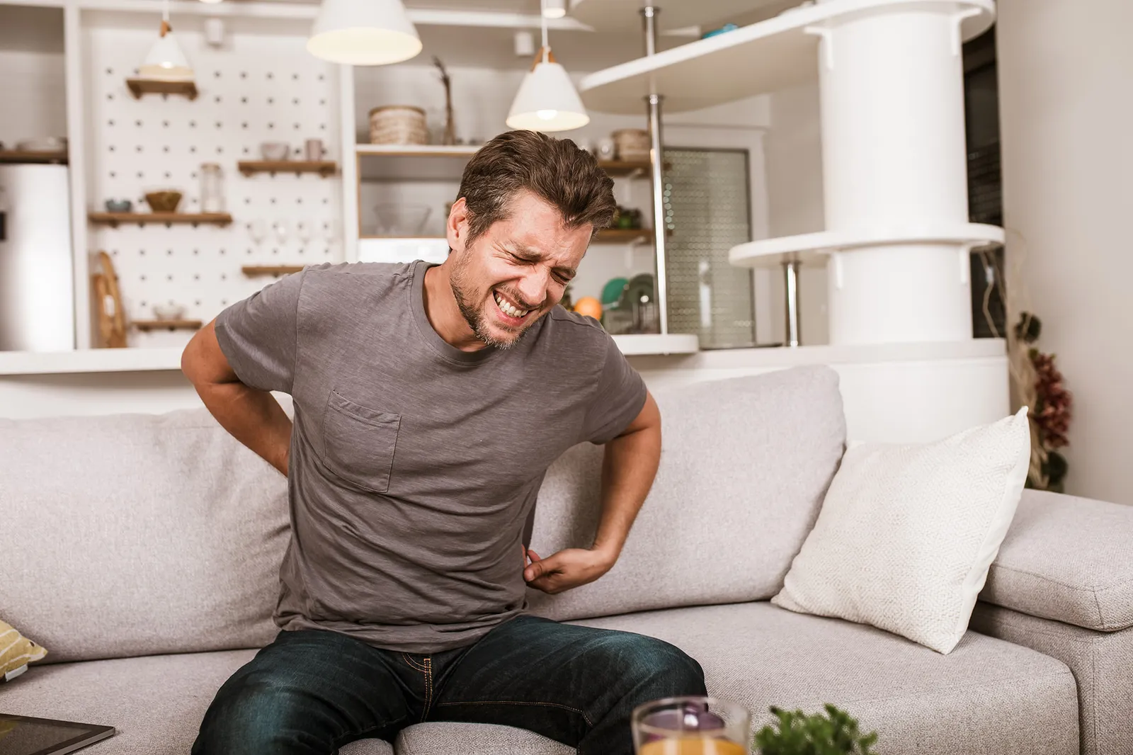 Ein Mann sitzt auf einem Sofa und hält sich vor Schmerzen den unteren Rücken. Sein Gesichtsausdruck zeigt, dass er unter starken Rückenschmerzen leidet.