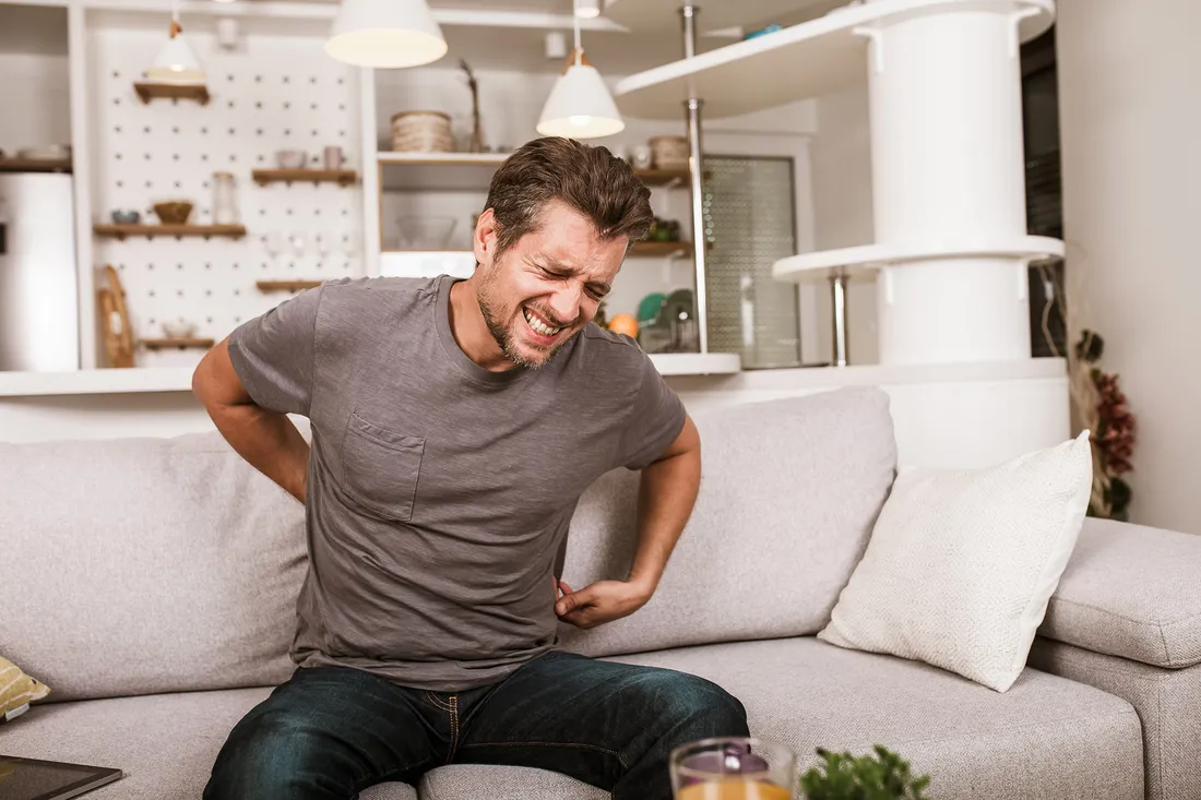 Ein Mann sitzt auf einem Sofa und hält sich vor Schmerzen den unteren Rücken. Sein Gesichtsausdruck zeigt, dass er unter starken Rückenschmerzen leidet.