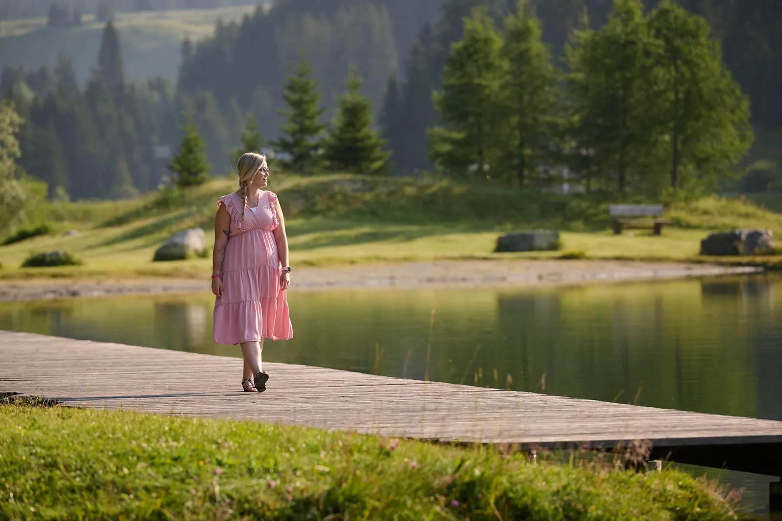 Eine Frau in einem rosa Kleid, die entspannt auf einem Holzweg an einem ruhigen See spaziert. Im Hintergrund sind grüne Wiesen und Bäume, die eine friedliche Atmosphäre schaffen.