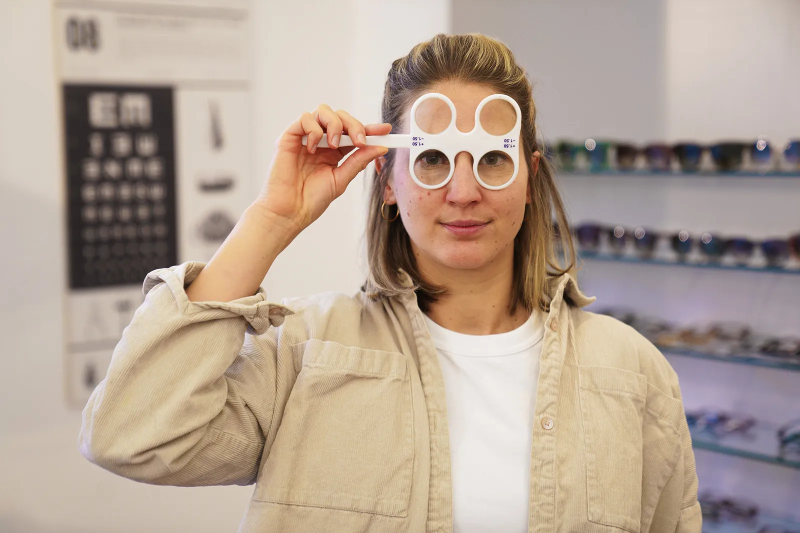 Eine Frau steht in einem Optikergeschäft und hält ein weisses Testgerät mit runden Löchern vor ihr Gesicht. Sie trägt eine helle Cordjacke und ein weisses T-Shirt, während im Hintergrund eine Wand mit Brillenmodellen sichtbar ist.