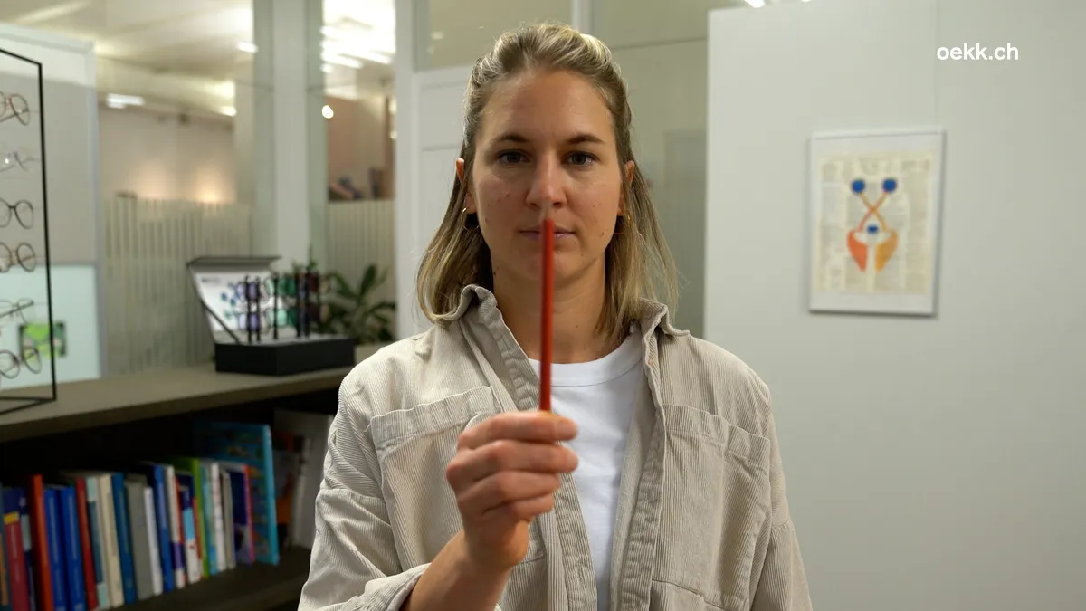 Frau führt eine Augenübung mit einem roten Stift durch, um die Augenmuskulatur zu stärken. Im Hintergrund sind Bücherregale und ein optisches Fachgeschäft zu sehen.