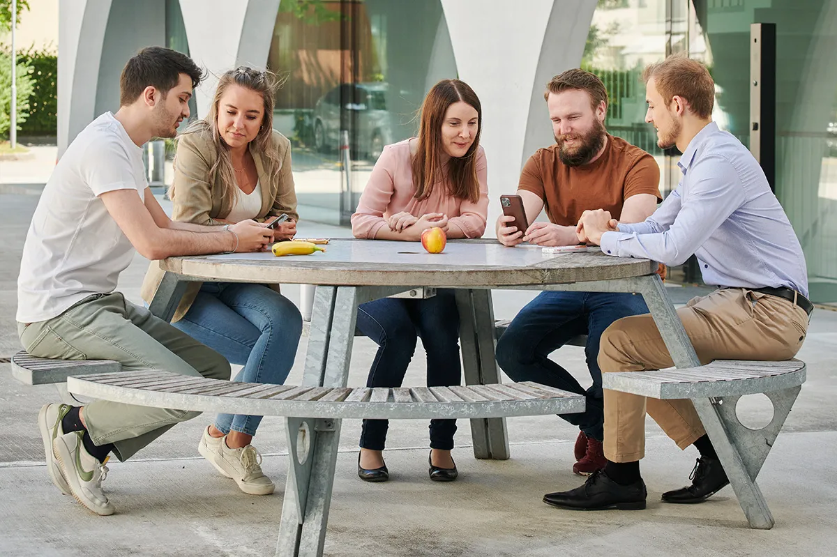 Cinque giovani adulti sono seduti insieme a un tavolo all’aperto, guardano uno smartphone e conversano in modo rilassato.