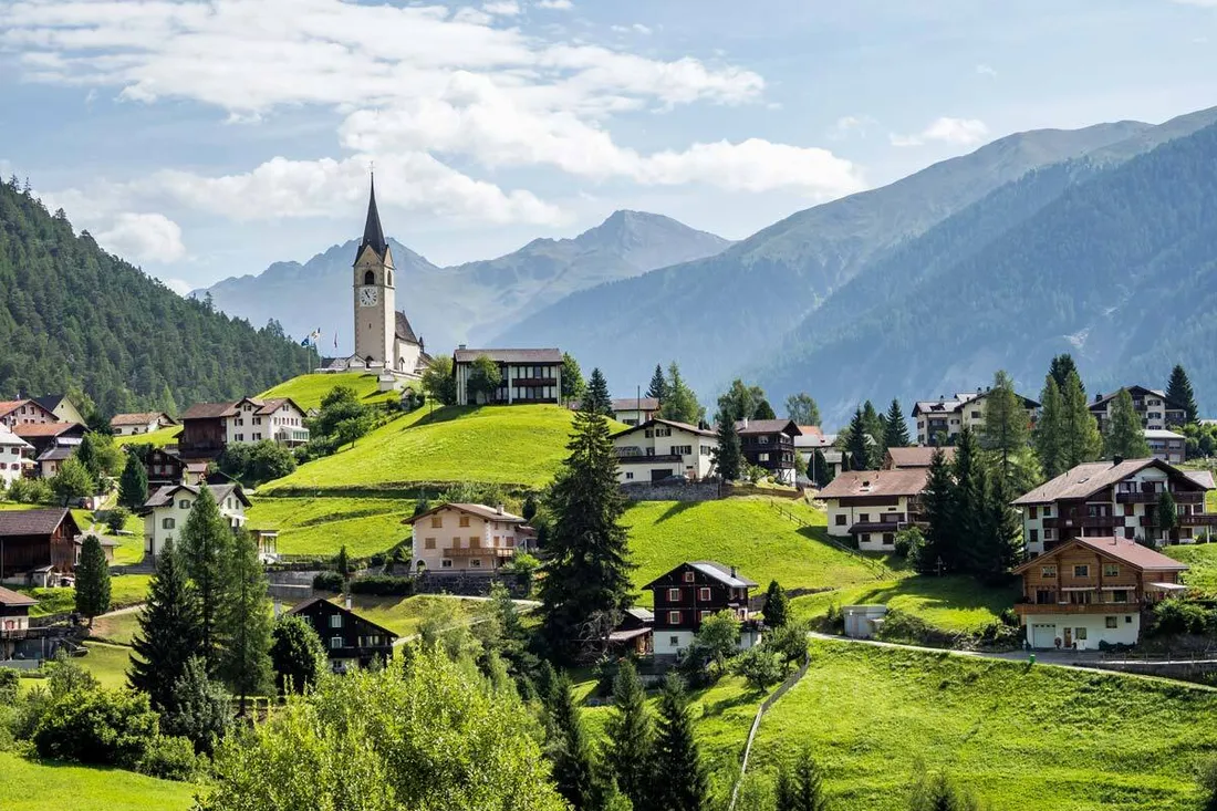 Un villaggio grigionese soleggiato con una chiesa su una collina, circondato da prati verdi, case tradizionali e un paesaggio alpino montano.