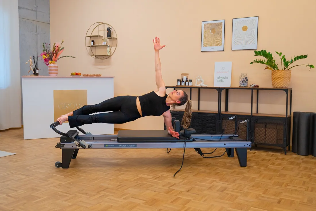 Frau in schwarzer Sportkleidung macht seitlichen Plank auf Pilates Reformer in hellem Raum mit Pflanzen und Wanddekoration