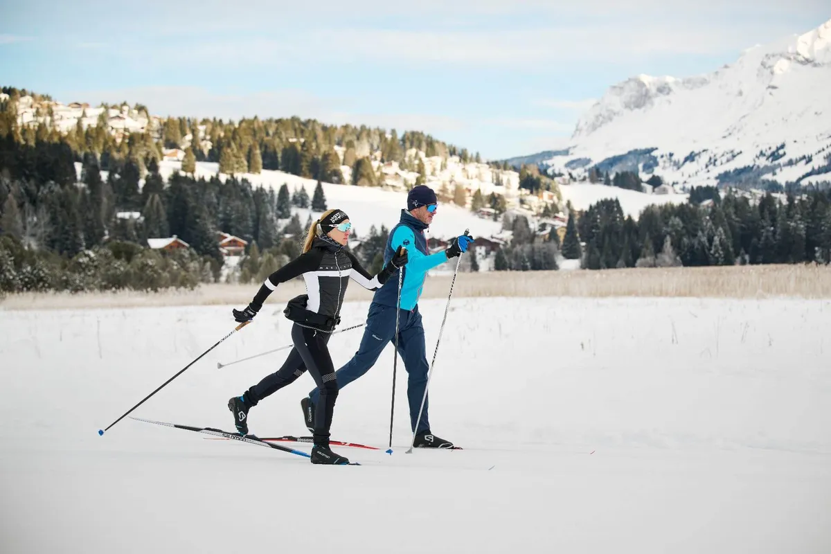 Due sciatori di fondo con abbigliamento invernale sciano fianco a fianco su una distesa innevata davanti a colline boschive e montagne.