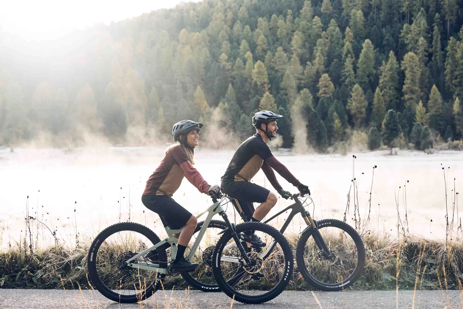 Zwei Personen fahren mit Mountainbikes entlang eines nebligen Sees vor bewaldeter Bergkulisse.