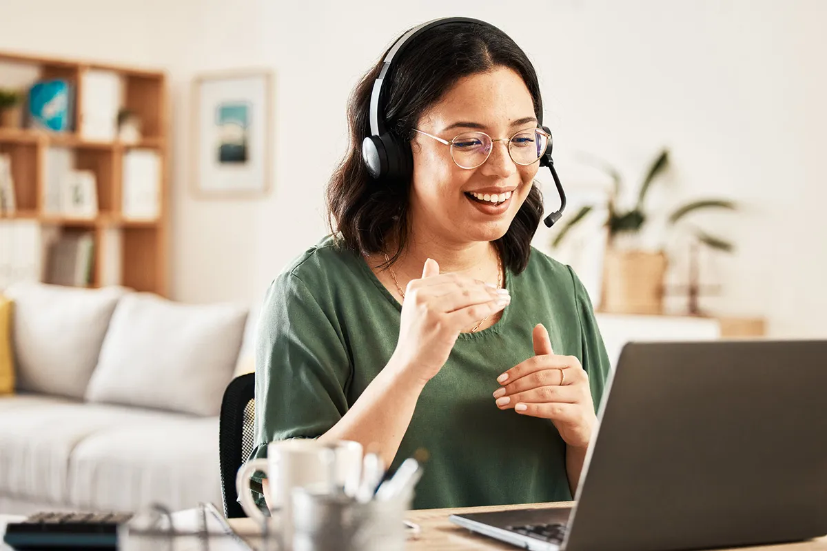 Una donna con cuffie e microfono è seduta davanti a un laptop a casa, sorride e spiega qualcosa gesticolando.