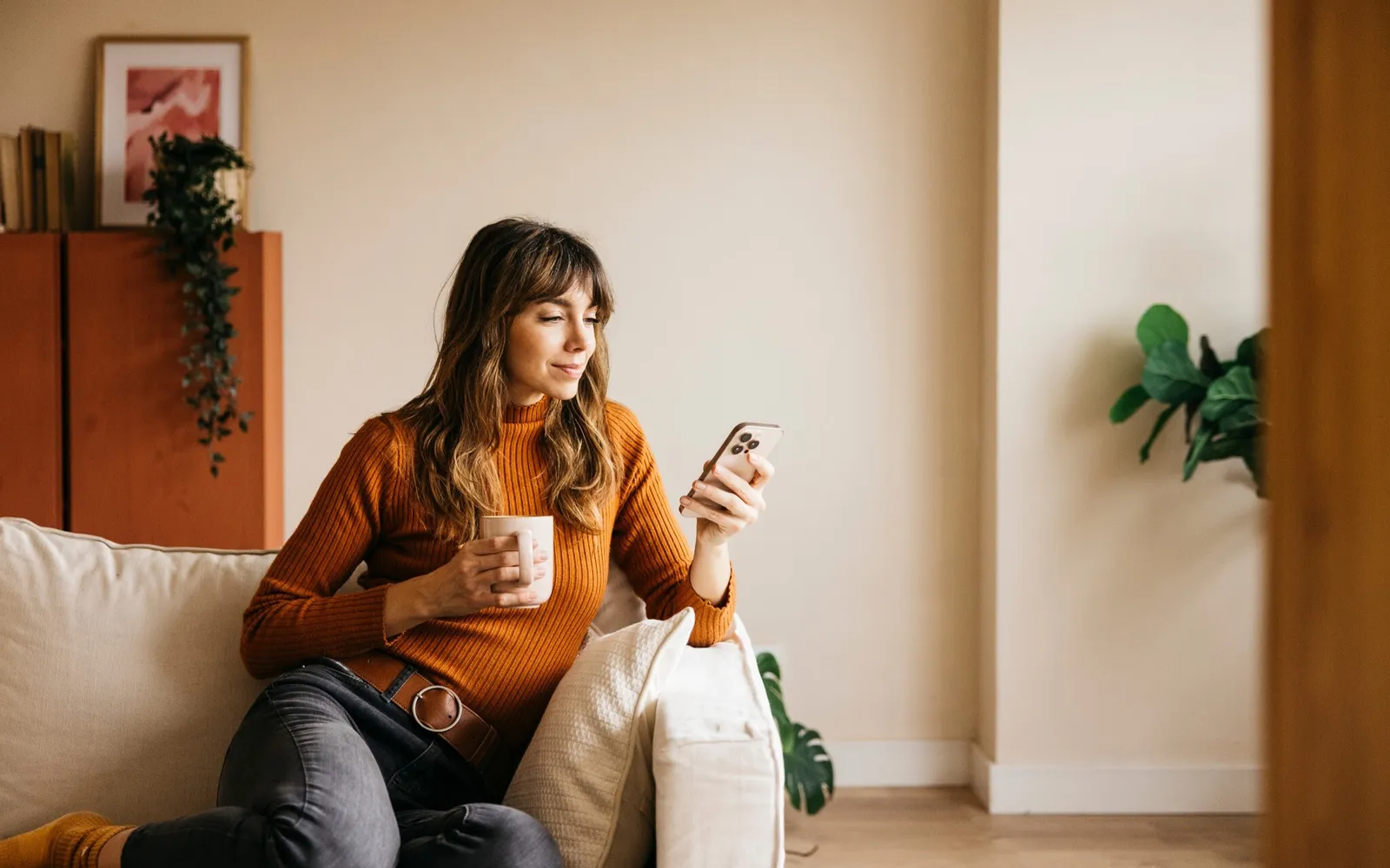 Una persona è seduta su un divano, tiene una tazza di caffè e guarda uno smartphone in un ambiente accogliente.