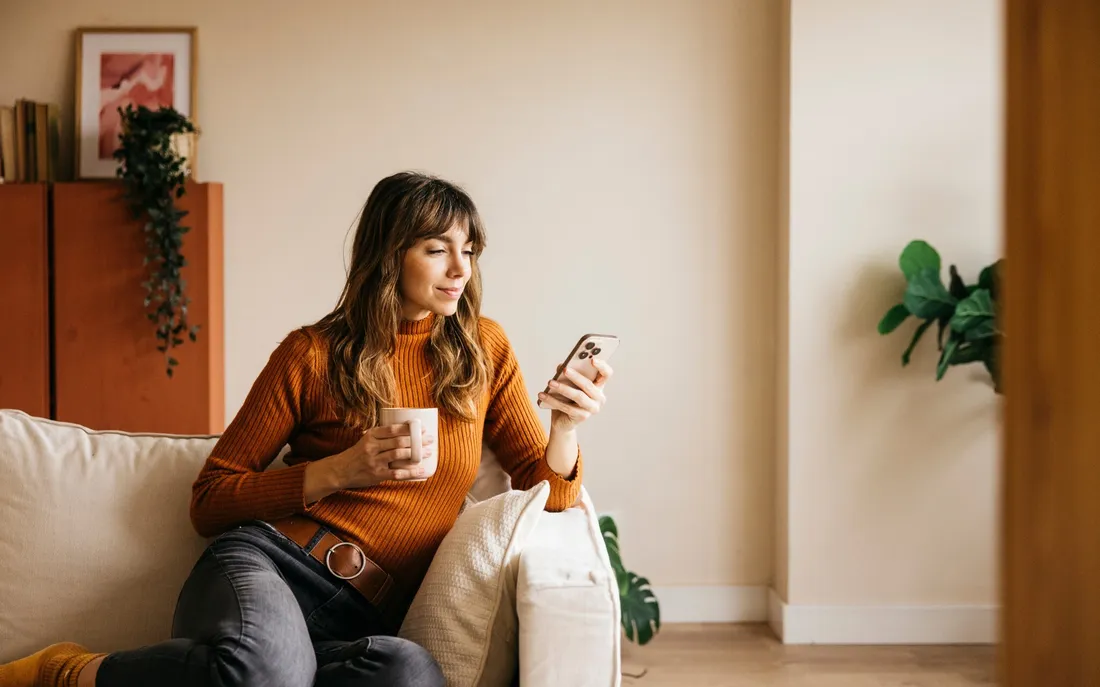 Una persona è seduta su un divano, tiene una tazza di caffè e guarda uno smartphone in un ambiente accogliente.