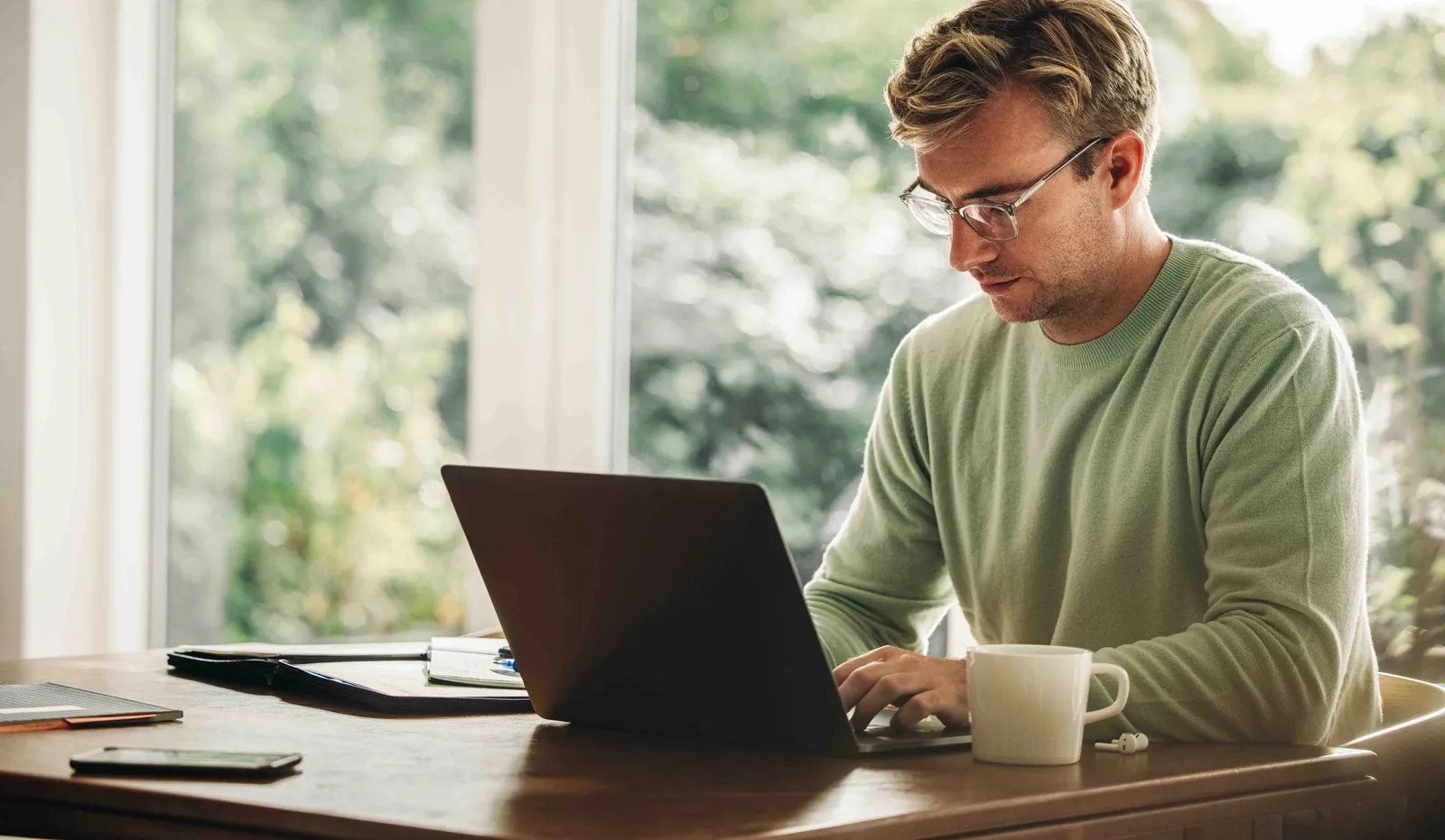 Personne travaille concentrée sur un ordinateur portable à une table avec café et notes, lumière naturelle en arrière-plan.