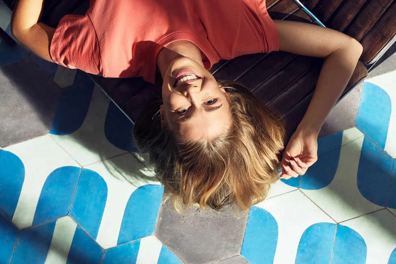 Persona con maglietta rosso-arancione sdraiata su un pavimento con piastrelle esagonali blu, bianche e grigie