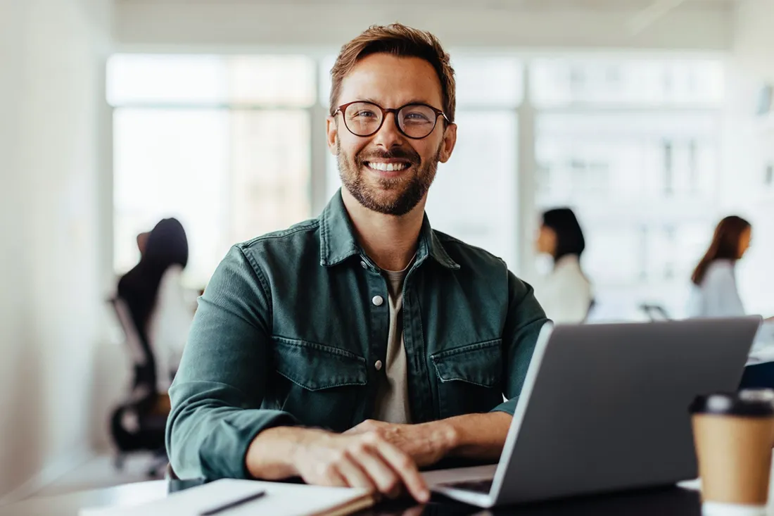 Uomo seduto a una scrivania con un laptop in un ufficio moderno con altre persone sullo sfondo