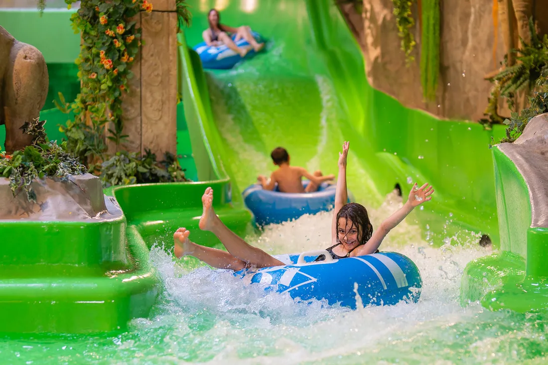 Ein fröhliches Kind rutscht auf einem blauen Gummireifen mit ausgestreckten Armen eine leuchtend grüne Wasserrutsche hinunter. Im Hintergrund sind weitere Kinder zu sehen, die ebenfalls die Rutsche genießen, umgeben von üppiger, dekorativer Bepflanzung und einer spielerischen Umgebung.